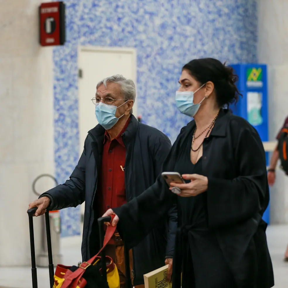 Caetano Veloso aparece com malas nas mãos e máscara de proteção no rosto. Paula Lavigne segura um celular com uma das mãos, enquanto leva as malas na outra