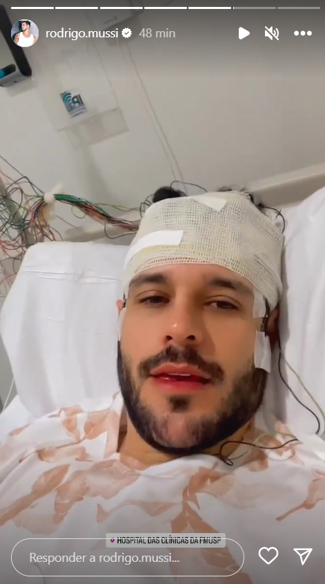 Rodrigo Mussi deitado em uma cama de hospital com a cabeça enfaixada e inúmeros fios ligados ao crânio