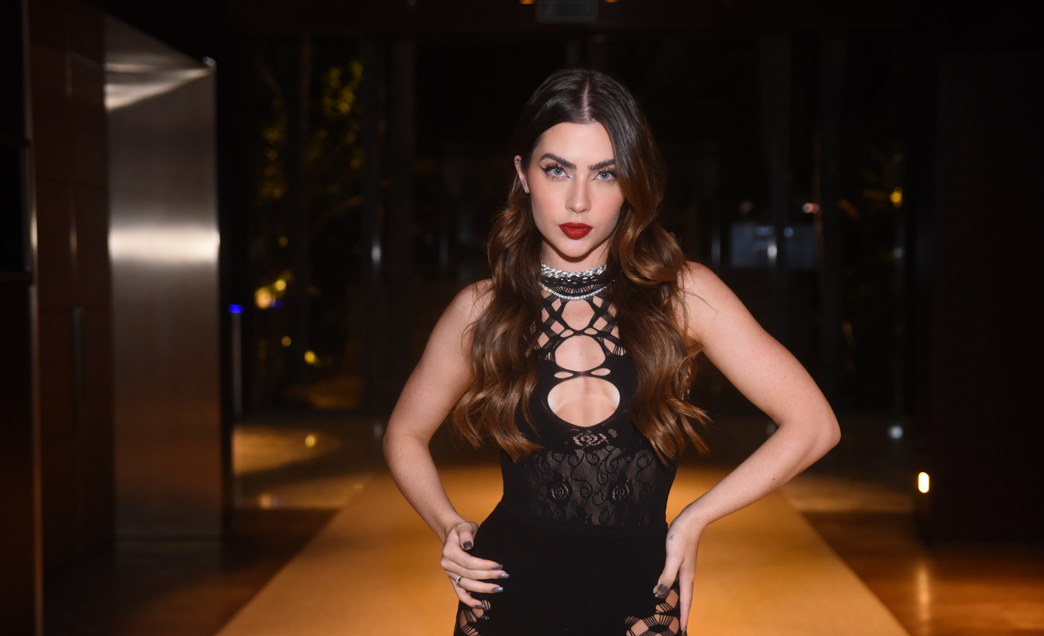 Foto de Jade Picon com um vestido preto cheio de recortes, batom vermelho e cabelo solto em uma entrada de festa