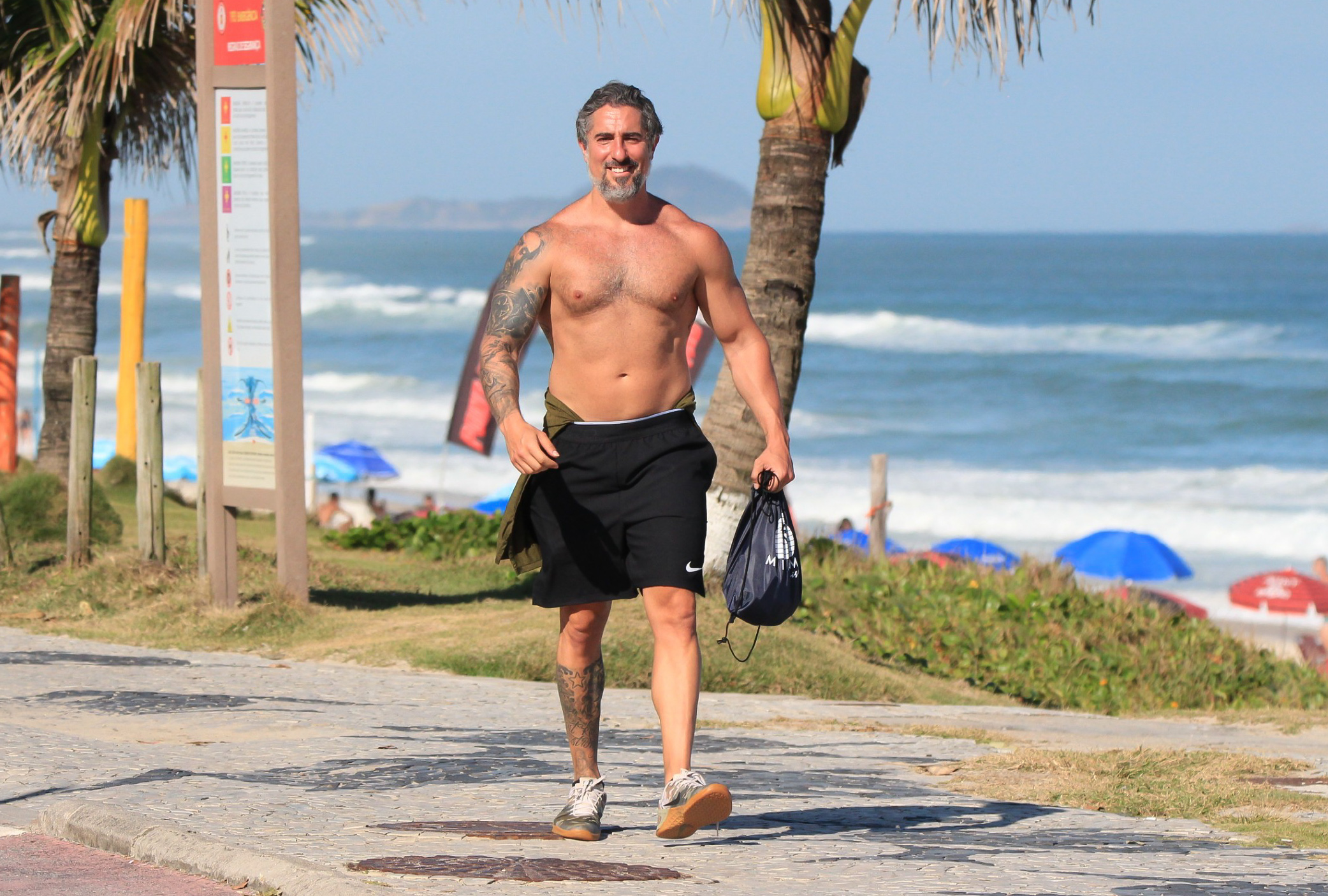 Foto de Marcos Mion andando sem camisa à beira da praia com corpo musculoso à mostra e sorridente