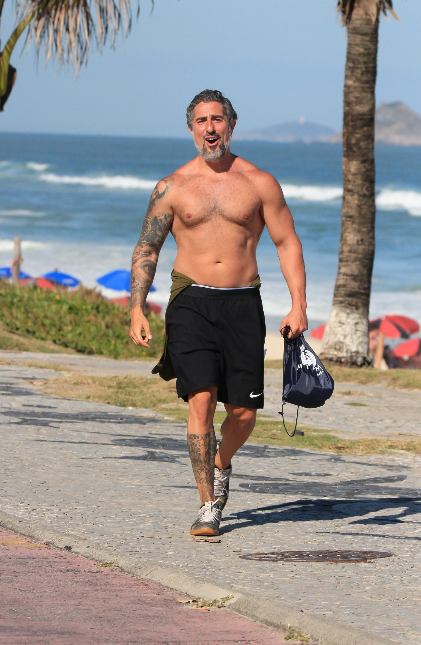 Foto de Marcos Mion andando sem camisa à beira da praia com corpo musculoso à mostra e sorridente