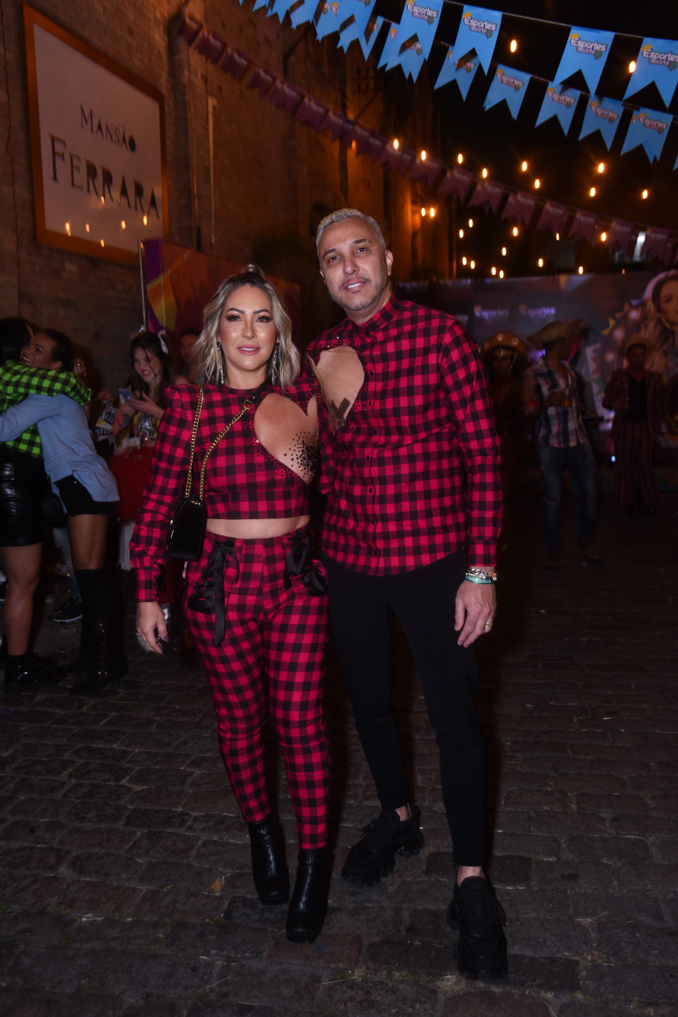 Foto de Claudia Baronesa e Rogério de camisa xadrez vermelha e preta, com recortes ao redor dos mamilos, cobertos por adesivos. Ao fundo, a festa junina Ressaca da Doutora