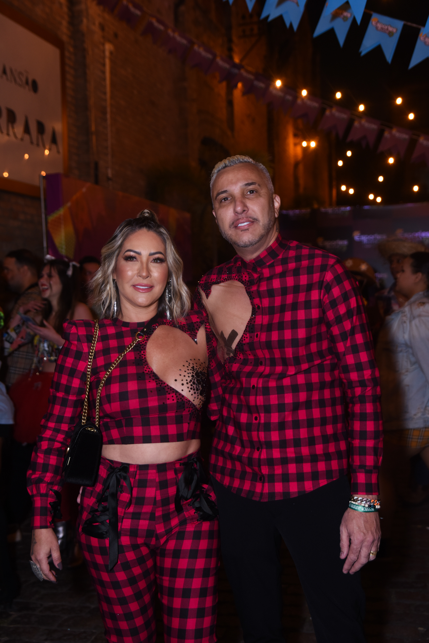 Foto de Claudia Baronesa e Rogério de camisa xadrez vermelha e preta, com recortes ao redor dos mamilos, cobertos por adesivos. Ao fundo, a festa junina Ressaca da Doutora