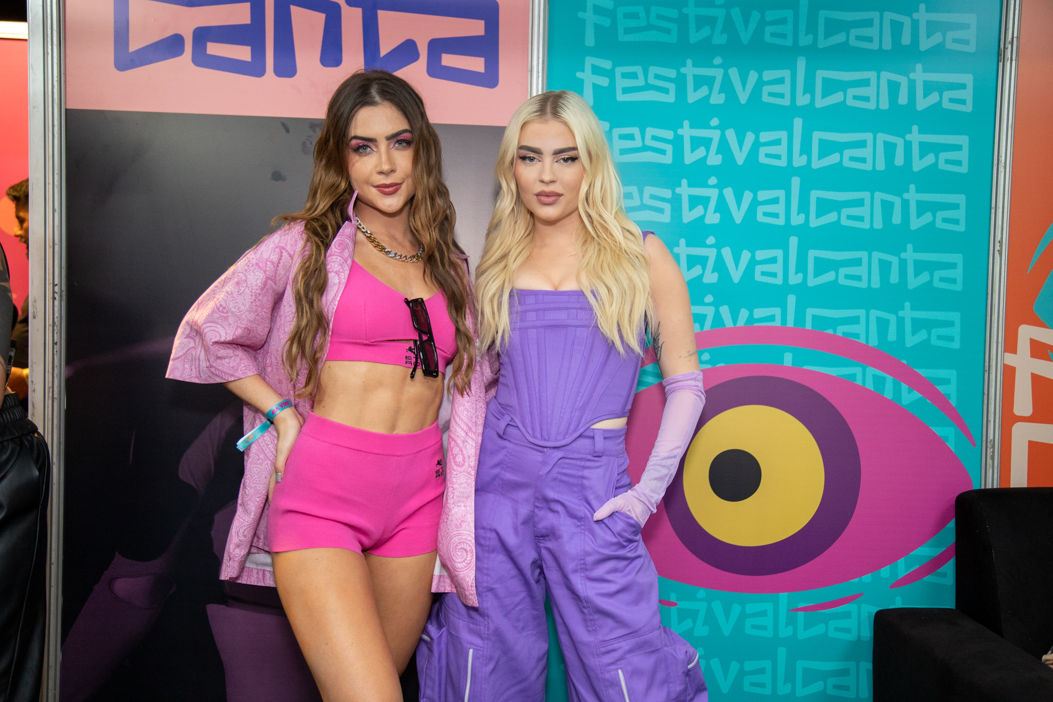 Jade Picon, de top e shortinho cor de rosa e um casaco aberto por cima, com os cabelos soltos e ondulados, posa ao lado de Luísa Sonza, com um conjunto de top e calça na cor roxa, em frente a um banner do festival Canta