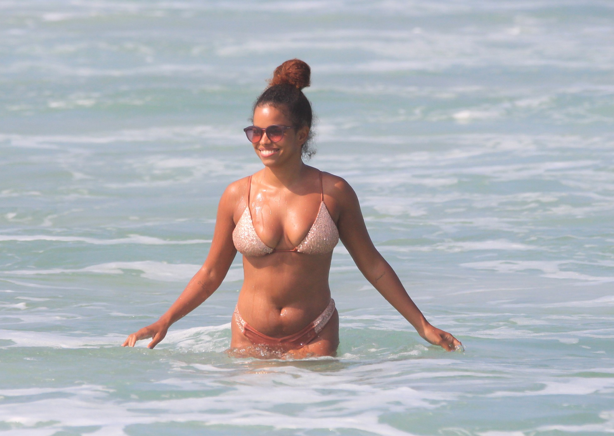 Foto de Jennifer Nascimento de biquíni de cordas finas em tom de barro e estampa branca, óculos escuros e cabelos presos, saindo da água do mar enquanto sorri