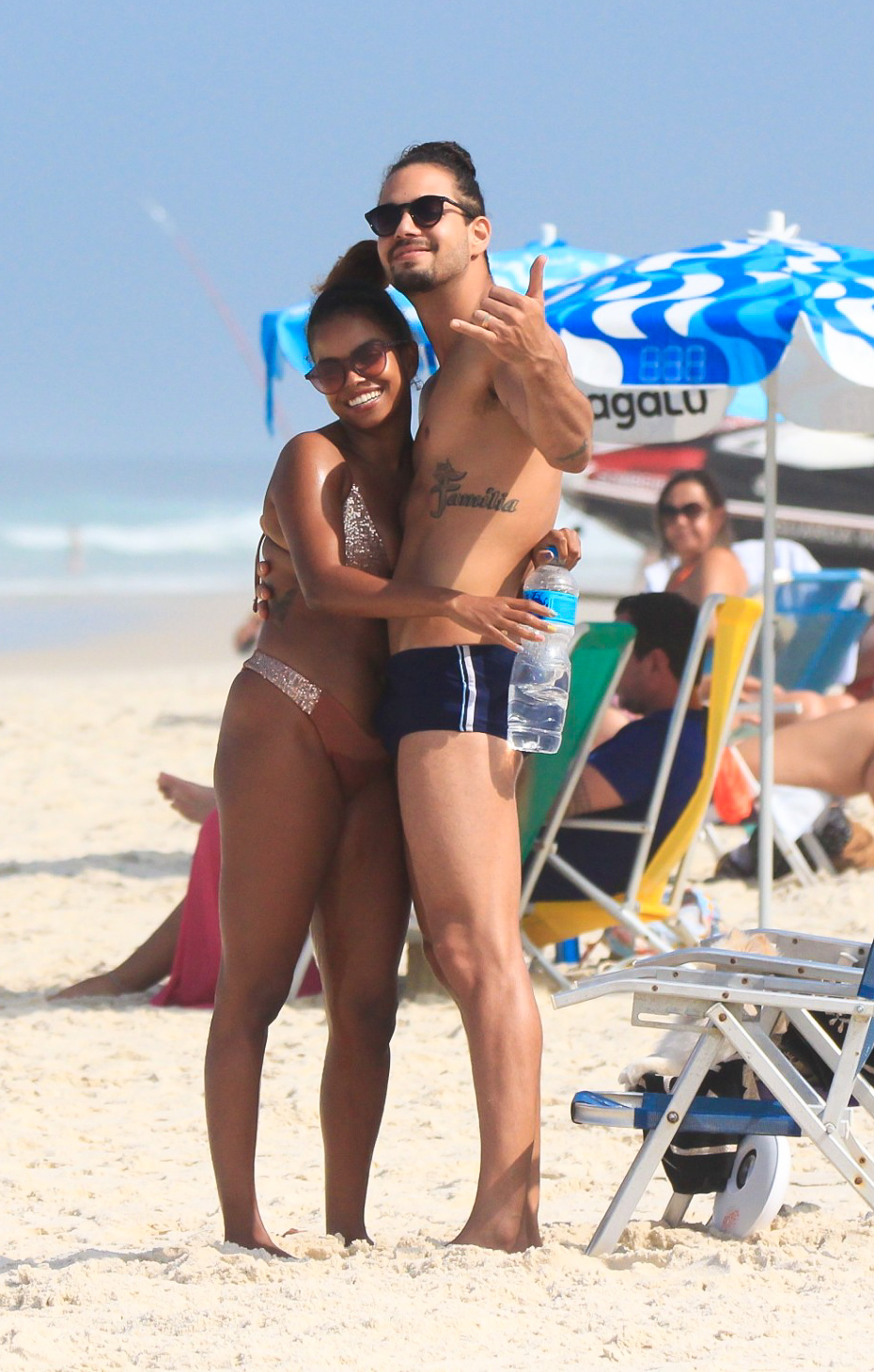 Foto de Jennifer Nascimento, de biquíni cor de terra e estampa branca, na praia, de cabelos presos e óculos escuros, segurando uma garrafa de água e abraça ao marido, o ator Jean Amorim, de sunga preta e branca e cabelos presos. Os dois olham e acenam para a câmera