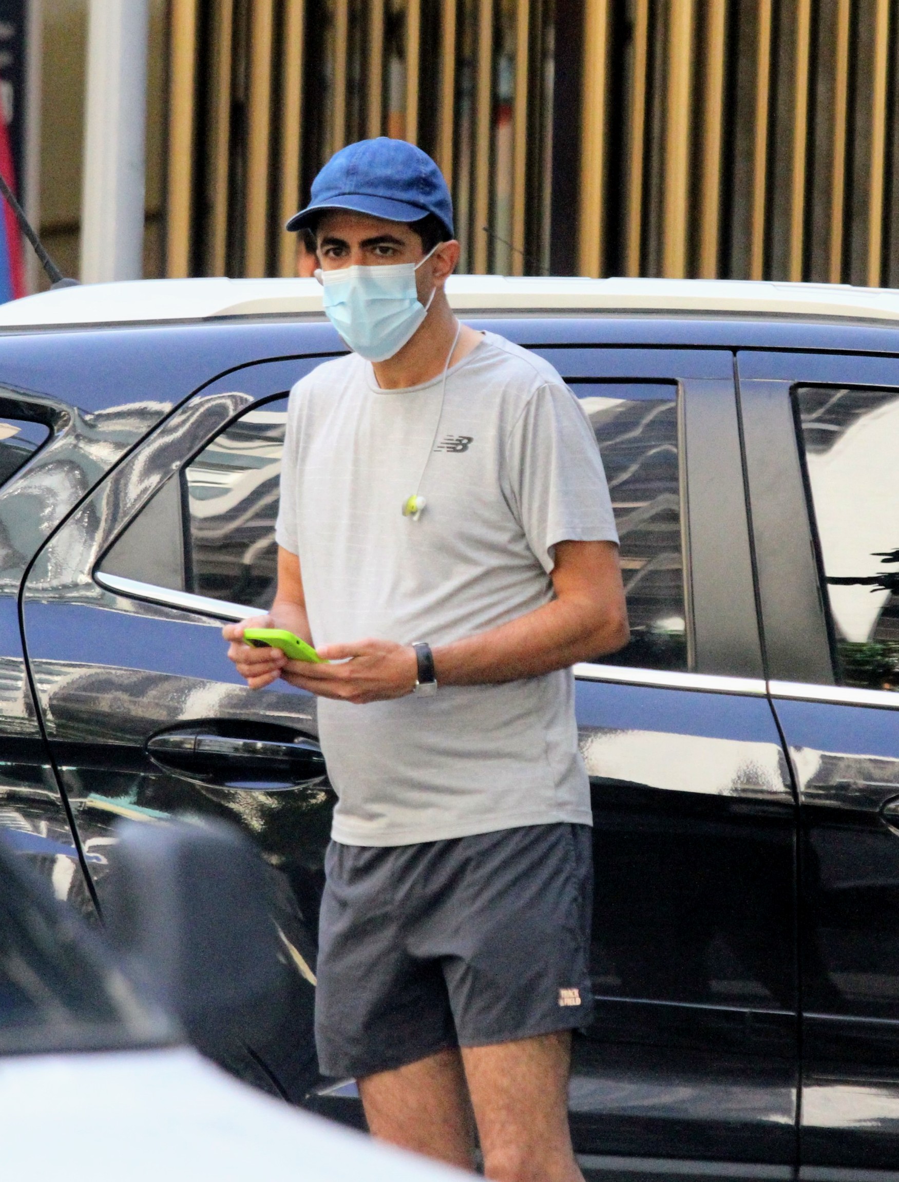 Foto de Marcius Melhem de boné azul, máscara cirúrgica, camiseta e bermuda segurando o celular