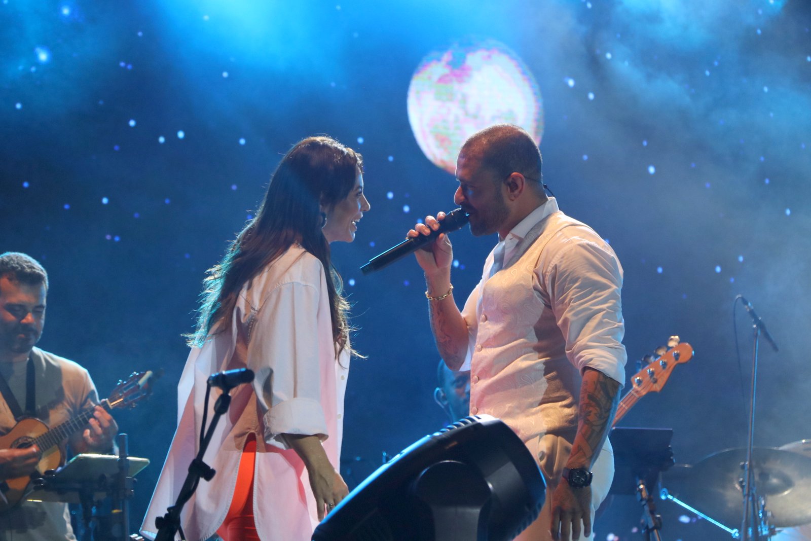 Foto de Paolla Oliveira e Diogo Nogueira no palco de um show, com um telão exibindo uma lua em um céu estrelado ao fundo enquanto o cantor canta no microfone e ela dança