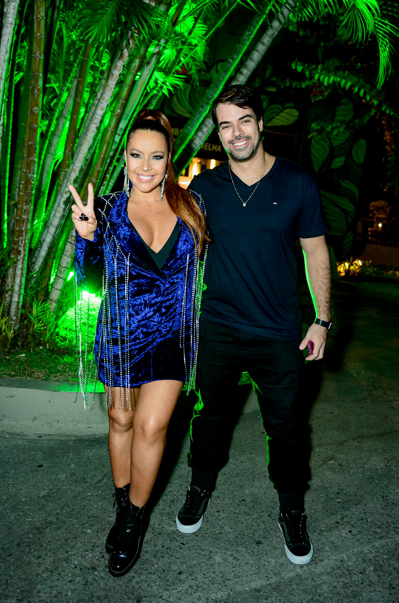 Foto de Renata Domingues em frente a plantas usando um vestido azul cintilante com detalhes em correntes prateadas e uma bota preta, com cabelos amarrados, ao lado do marido, Leandro Gleria