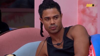 Participante do BBB 23, Gabriel Santana, usa um top preto e tem tranças no cabelo, sentado em um sofá rosa.
