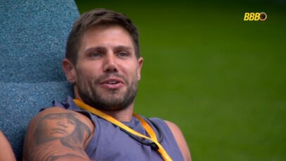 Homem com tatuagem no braço, sentado em poltrona azul, com logo do BBB ao fundo.