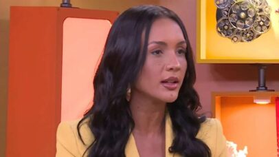 A mulher com cabelos longos e escuros e um blazer amarelo, em um estúdio de TV com decoração laranja e engrenagens ao fundo.