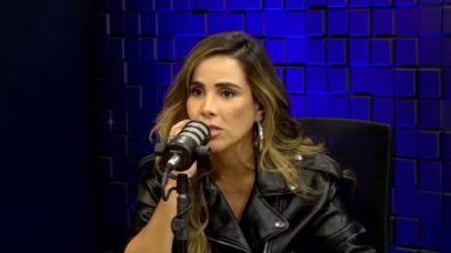 A mulher de cabelos castanhos, usando jaqueta de couro preta, fala em um microfone em um estúdio com fundo azul.